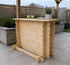 A light natural wood outdoor bar stool with a rustic, stacked-log design, set on a paved patio with a wooden fence and garden plants in the background.