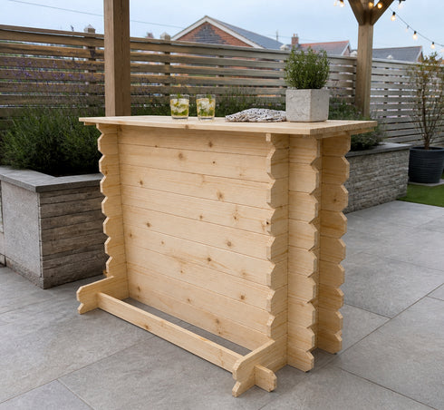 A light natural wood outdoor bar stool with a rustic, stacked-log design, set on a paved patio with a wooden fence and garden plants in the background.