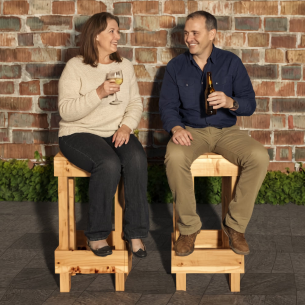 Two people sitting on wooden stools against a brick wall, one holding a glass of wine and the other a bottle.